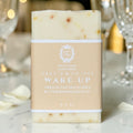 A bar of Wake Up handmade soap by Oracie & Co 1942, wrapped in a beige paper label, standing on a marble surface with a softly blurred background of glassware and warm lights.