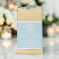 A single bar of handmade soap standing upright on a light marble surface with a softly blurred indoor background and warm bokeh lights. The soap has a pale cream body with a light blue paper label wrapped around the middle.
