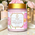 A jar of Gracie & Co 1942 Pink Peony Nourishing Sugar Scrub, featuring a white label with pink peony illustrations and a metallic gold lid, set against a soft-focus background of pink flowers and warm lights.